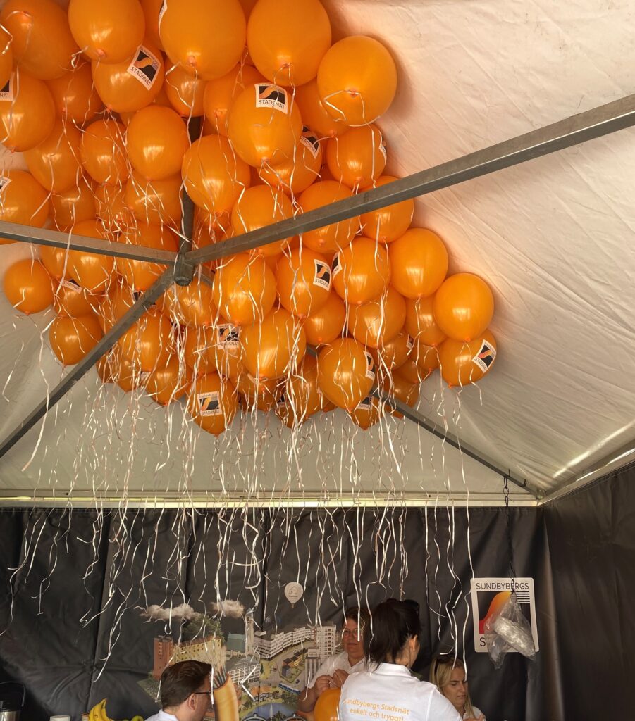 Massor av upplåsta orange heliumballonger med Sundbybergs Stadsnäts logotyp hänger ned från taket i ett tält. Ballongsnörena hänger ned och fyra personer sitter ned och fyller ballonger med gas. 
