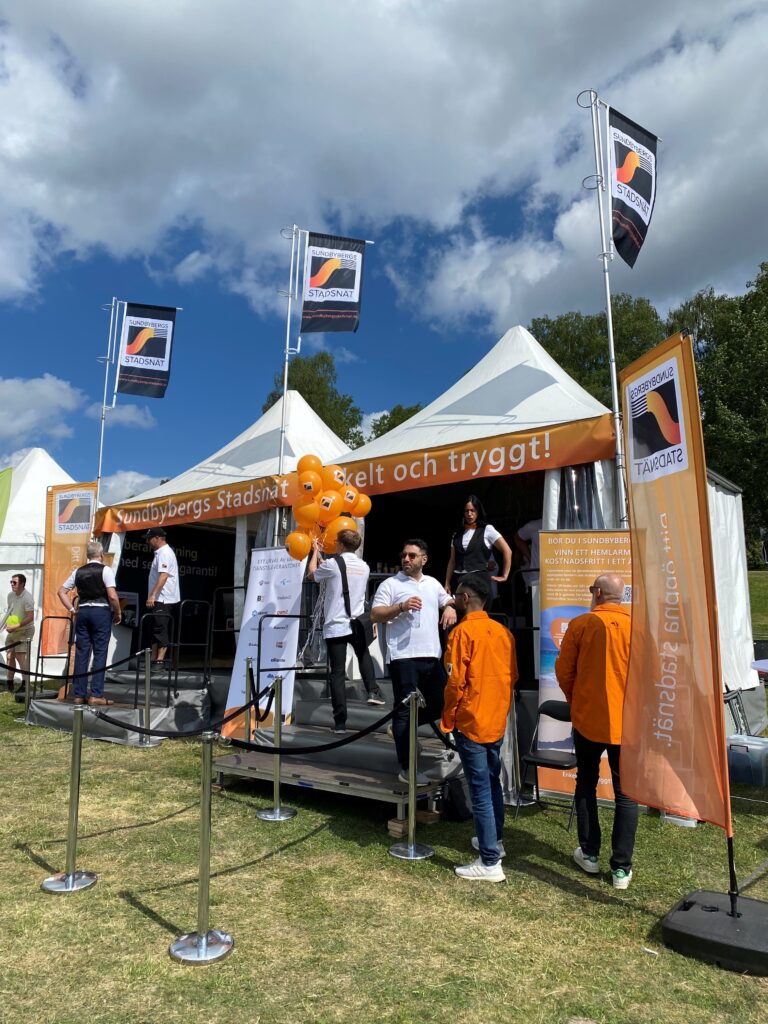 Människor klädda i orange och vita kläder står utanför ett tält som pryds med flaggor som det står Sundbybergs Stadsnät på.